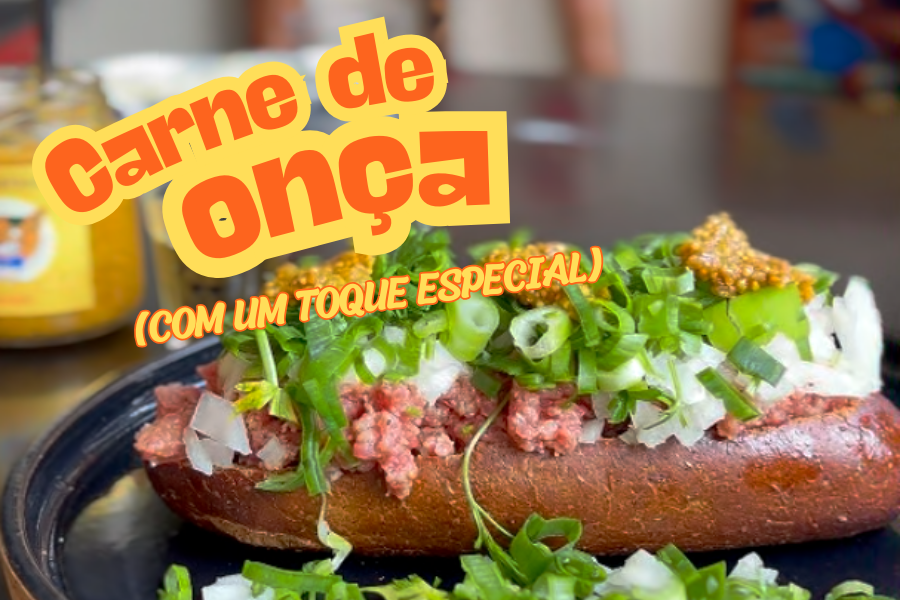 Imagem de um prato de carne de onça, constituído por um pão escuro, carne crua moída e temperada, coberta com cebola picada, cebolinha e salsinha, finalizado com mostarda da Onça Bêbada. Ao fundo, um copo de dose com cachaça e um pode de mostarda da Onça Bêbada