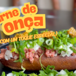 Imagem de um prato de carne de onça, constituído por um pão escuro, carne crua moída e temperada, coberta com cebola picada, cebolinha e salsinha, finalizado com mostarda da Onça Bêbada. Ao fundo, um copo de dose com cachaça e um pode de mostarda da Onça Bêbada